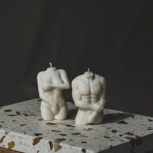 Two sculptural candles designed to resemble human figures, placed on a surface with a marble-like texture.